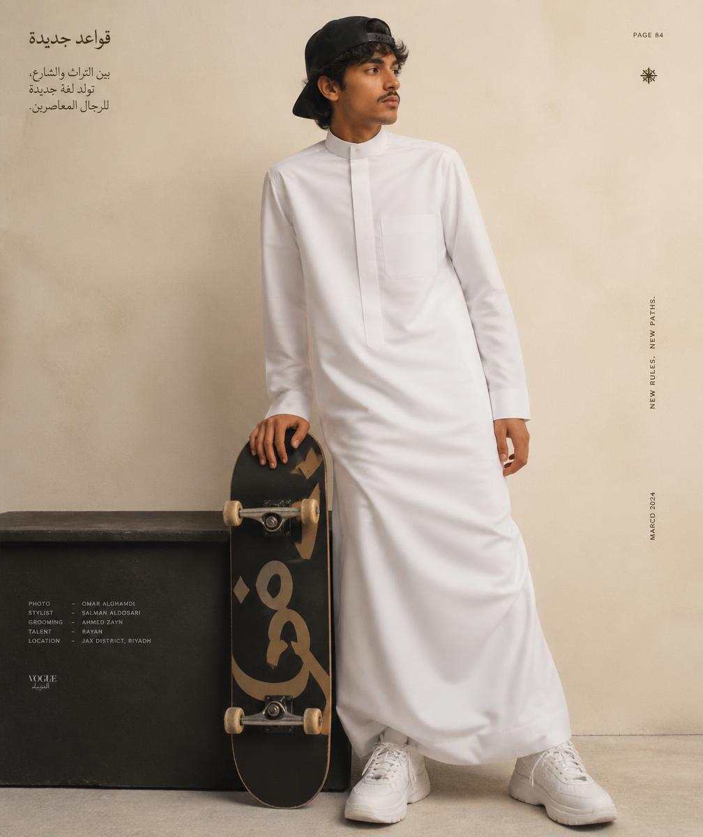 A young Saudi man in a white thobe, backwards cap and chunky sneakers resting on a skate deck